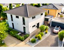 Moderne zweigeschossige Doppelhaushälfte mit weißer Fassade und dunklem Satteldach, umgeben von einem gepflasterten Hof, einem kleinen Vorgarten mit Ziergräsern und einer Einfahrt mit geparktem Auto. Im Hintergrund sind Nachbarhäuser und grüne Bäume zu sehen.