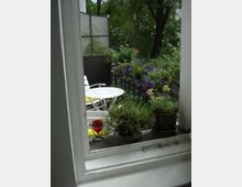 Blick durch ein geöffnetes Fenster auf einen kleinen Balkon mit Pflanzen und Blumen. Es sind ein runder weißer Tisch, zwei weiße Stühle und ein schmiedeeisernes Geländer zu sehen, im Hintergrund Bäume und grüne Vegetation.