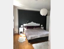 Schlafzimmer mit einem großen Bett, das eine verzierte weiße Kopfteilleiste hat, vor einer dunkel gestrichenen Wand. Auf beiden Seiten befinden sich weiße Nachttische, eine Feder-Pendelleuchte hängt von der Decke, und der Boden ist aus hellen Holzdielen mit weißen Fellteppichen.
