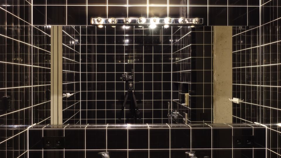 Badezimmer mit schwarzen, glänzenden Fliesen an Wänden und Decke, einem weißen rechteckigen Waschbecken, und einem Spiegel mit integrierter Beleuchtung. Die Armaturen sind schlicht, und der Raum wird durch künstliches Licht erhellt.