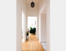 Ein lichtdurchfluteter Flur mit Holzboden führt zu einem hellen Wohnzimmer, in dem ein grünes Sofa und Pflanzen stehen. Eine schwarze Deckenleuchte und ein Pflanzenständer mit einer Topfpflanze zieren den schlichten, weißen Raum.