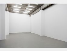 Leerer, rechteckiger Raum mit weißen Wänden und dunkelgrauem Betonboden. Die Decke besteht aus sichtbaren Betonbalken mit parallel verlaufenden Leuchtstofflampen.