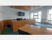 Ein moderner Gerichtssaal mit Holzmöbeln, darunter ein Richterpult und Tische für andere Beteiligte. Große Fenster lassen Tageslicht herein, und an der Wand hängt ein Flachbildschirm.