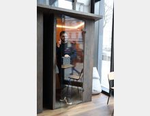 Ein hölzernes, schallisoliertes Telefonhäuschen mit einer Glastür, in dem ein Mann telefoniert. Das Häuschen steht in einem modernen Raum mit großen Fenstern und Holzboden, neben einer Sitzgelegenheit.