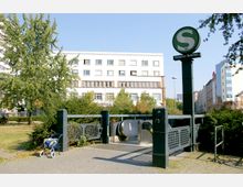 Eingang zur S-Bahn-Station "Anhalter Bahnhof" in einer städtischen Umgebung, mit einem grünen Schild mit S-Bahn-Logo, einer Rolltreppe und umliegender Vegetation im Vordergrund. Im Hintergrund sind ein mehrstöckiges weißes Gebäude und weitere städtische Strukturen sichtbar.
