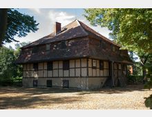 Fachwerkhaus mit schrägem Ziegeldach und gemauertem Sockel, umgeben von Bäumen. Das Gebäude steht auf einem mit Laub bedeckten Grundstück und hat einen kleinen Eingang mit Holztreppe auf der rechten Seite.