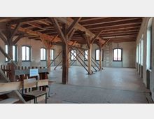 Ein leerer Dachboden mit sichtbarer Holzbalkenkonstruktion, schrägen Stützbalken und hellem Holzfußboden. Die Wände bestehen aus unverputztem Ziegelstein und verfügen über mehrere hohe Bogenfenster, die Tageslicht hereinlassen.