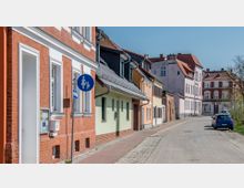 Straßenszene in einer Kleinstadt mit Reihen von Wohnhäusern aus Ziegel- und Putzfassaden in verschiedenen Farben. Der Bürgersteig ist mit Kopfsteinpflaster gestaltet, auf der Straße parken Autos, und ein Fußgängerschild ist sichtbar.