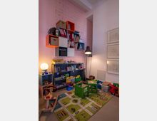 Spielzimmer mit einer rosa Ziegelwand, Regalen voller Spielzeug, Büchern und Ordnern sowie einer grünen Spielmatte mit Straßenmotiven. Im Raum befinden sich ein kleiner Holztisch mit zwei Stühlen, eine Lampe und verschiedene Kinderspielzeuge, darunter ein Schaukelpferd und Bauklötze.