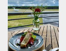 Ein Holztisch mit einem Teller, darauf ein Gericht aus Fischfilet, Salat und roten Beilagen, steht auf einer Terrasse mit Blick auf eine Landebahn. Im Hintergrund sind ein kleines Flugzeug sowie Bäume zu sehen, daneben eine Vase mit roten und weißen Blumen.
