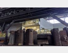 Industriehalle mit einer Mischung aus alten Stahlbauteilen im Vordergrund und modernen Elementen wie einer verglasten Galerie mit Geländern und Treppe im Hintergrund. Große Fenster und Stahlträger prägen die Architektur.