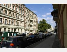 Straßenszene in einem Wohngebiet mit mehrstöckigen Altbauhäusern im Stil des 19. Jahrhunderts, verziert mit roten Backsteinelementen und weißen Putzverzierungen. Autos sind am Straßenrand geparkt, und im Hintergrund sind Bäume und weitere Häuser zu sehen.