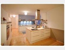 Moderne Küche mit hellen Holzböden, einer Kücheninsel mit integriertem Kochfeld und weißen Schränken. Im Hintergrund blaue Wand mit Arbeitsfläche, Spüle und offenen Regalen.