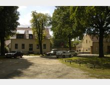 Wohnhäuser in ländlicher Umgebung mit hellen Fassaden und roten Ziegeldächern, umgeben von hohen Bäumen. Im Vordergrund stehen mehrere geparkte Autos auf einem unbefestigten Platz, umrahmt von Grünflächen.