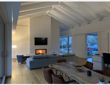 Modernes Wohnzimmer mit hohen, weißen Holzbalkendecken, einem Kamin unter einem Wandfernseher, einer großen grauen Couch und einer offenen Esszimmer-Ecke mit einem Holztisch. Große Glasfenster bieten Blick auf einen Garten und geparkte Autos.