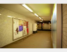 Ein U-Bahn-Tunnel mit hell beleuchteten weißen Fliesenwänden, Werbetafeln auf der linken Seite, einem Fahrkartenautomaten rechts und einem leicht beleuchteten Bahnsteig im Hintergrund. Die Decke ist mit langen Leuchtstofflampen ausgestattet.