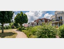 Mehrfamilienhäuser mit Balkonen und roten Schrägdächern in einer grünen Wohnsiedlung; ein geschwungener Kiesweg führt durch eine parkähnliche Anlage mit Bäumen, Sträuchern und Rasen unter einem wolkigen Himmel.