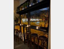 Eine gemütliche Café-Ecke mit hohen Barhockern und einer schwarzen Theke vor großen Fenstern, die nachts auf eine beleuchtete Straße mit einem Supermarkt und Bäumen blicken; Kerzen und Zimmerpflanzen dekorieren den Raum.