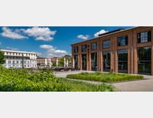 Backstein-Gebäude mit großen, rechteckigen Fenstern auf der rechten Seite, umgeben von Grünflächen und einem gepflasterten Weg; im Hintergrund ein Platz mit weißen Sonnenschirmen und weiteren Gebäuden unter blauem Himmel mit Wolken.