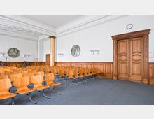 Ein heller Raum mit mehreren Reihen hölzerner Stühle, einer Tür mit aufwendigen Holzvertäfelungen, einer Säule und Wandleuchten. Die Wand ist weiß mit zwei runden Wandreliefs und einer Uhr oberhalb der Tür.