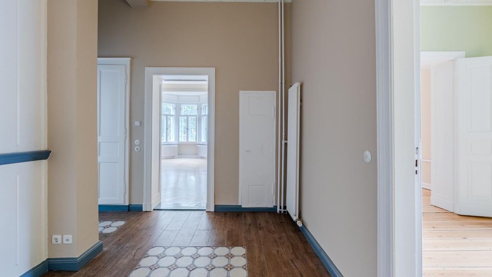 Heller Flur in einer Altbauwohnung mit hohen Decken und Stuck, beige gestrichenen Wänden, weißen Türrahmen und blau abgesetzten Sockelleisten. Dunkler Holzboden mit einem Abschnitt aus gemusterten Fliesen; mehrere Türen führen in weitere Räume, am Ende ist ein Raum mit großen Fenstern sichtbar.
