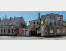 Straßenansicht eines verlassenen, verfallenen Gebäudekomplexes mit teilweise freiliegendem Mauerwerk, zerstörten Fenstern und einem Schornstein im Hintergrund. Ein Schild am Eingang weist auf ein Atelier hin, daneben steht ein geschlossenes Tor.