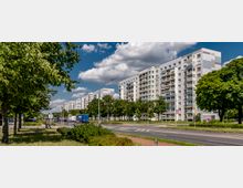 Mehrstöckige Wohngebäude mit Balkonen entlang einer Straße, umgeben von Grünflächen und Bäumen. Der Himmel ist blau mit vereinzelten Wolken, und am Straßenrand sind Fahrzeuge und Fußgänger zu sehen.