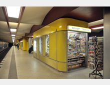 U-Bahn-Station mit gelb verkleidetem Kiosk, der Zeitungen, Zeitschriften und Tabakwaren verkauft. Im Hintergrund sind ein Bahnsteig, Sitzbänke und wenige Passanten zu sehen.