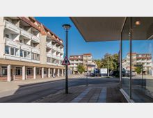 Straßenszene mit mehrstöckigen Wohngebäuden mit weißen Balkonen und roten Dächern, einem Gehweg mit Beleuchtung und einem Verkehrsschild in der Mitte des Bildes. Im Hintergrund stehen weitere Gebäude und Bäume, während rechts im Vordergrund eine Glasfassade sichtbar ist.