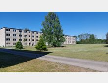 Mehrstöckiges, schlichtes Plattenbaugebäude in einem grasbedeckten, offenen Bereich mit vereinzelten Bäumen und einem kleinen Fußballtor. Im Vordergrund verläuft ein geteilter Gehweg, umgeben von Rasenflächen.