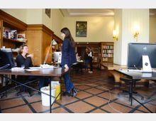 Großraumbüro mit klassischem Interieur: Holzwände, eingebaute Regale voller Bücher und mehrere Tische mit Computern. Im Vordergrund befinden sich zwei Personen an einem Schreibtisch, während im Hintergrund weitere Leute zusammenarbeiten.