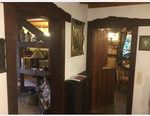 Ein rustikaler Wohnbereich mit Holzbalken, einer halboffenen Holztür und antiken Dekorationsgegenständen wie Puppen, Keramiken und einem Bild an der Wand. Durch die Durchgänge sind eine einladende Sitzecke, eine Lampe und ein Fenster mit Gardinen sichtbar.