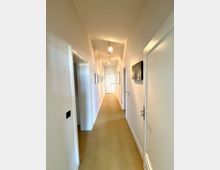 Langer, schmaler Flur in einer Wohnung mit weißen Wänden, beige-farbenem Boden und mehreren Türen auf beiden Seiten. An der Decke hängen einfache Leuchten, und an den Wänden sind gerahmte Bilder sowie ein kleines Wandregal angebracht; am Ende des Flurs ist eine geschlossene Tür sichtbar.