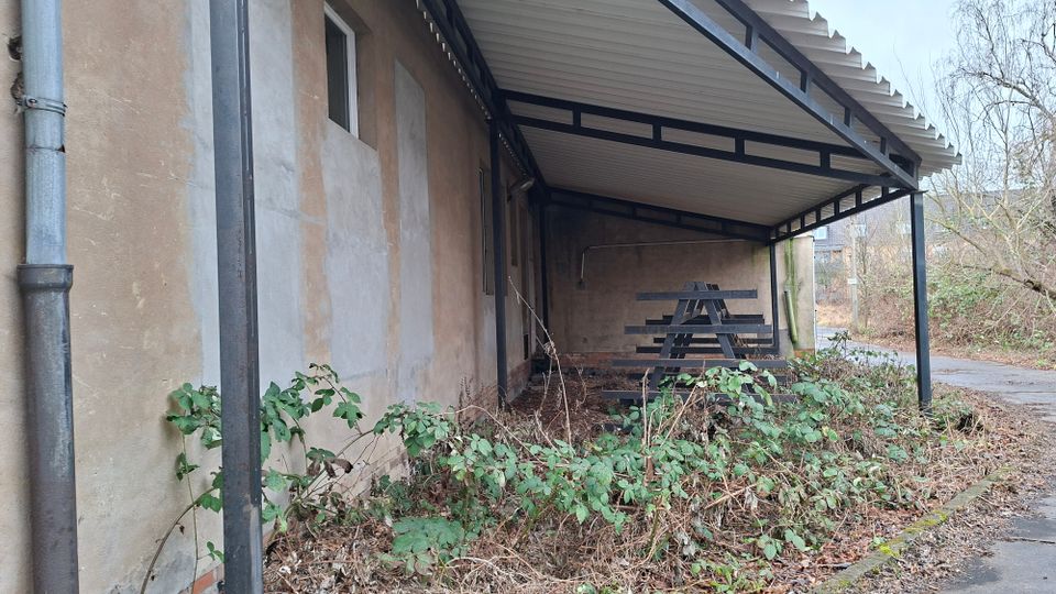 Überdachter Anbau an einem verputzten Gebäude mit Metallstützen und Wellblechdach; darunter steht eine lange, dunkle Sitzbank. Im Vordergrund wuchert Gestrüpp und Laub, daneben verläuft ein schmaler Weg am Rand eines baumreichen Grundstücks.