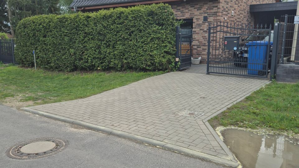 Einfamilienhaus aus Backstein mit steilem, dunkel gedecktem Satteldach und zwei kleinen Fenstern im Giebel. Davor liegen eine gepflasterte Einfahrt mit Metalltor, hohe Hecke und ein kleiner Vorgarten entlang einer Wohnstraße.
