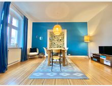 Wohnzimmer mit Holzdielenboden, einem Esstisch, umgeben von modernen Stühlen, und einer blauen Akzentwand. Links sind große Fenster mit blauen Vorhängen, rechts ein Fernseher auf einem Holzregal und eine Stehlampe, im Hintergrund eine Tapete mit Pflanzenmuster.