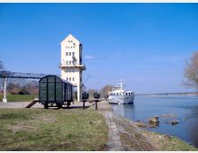 Ein Flusshafen mit einem markanten weißen Turmgebäude, das sich nahe am Ufer befindet. Im Vordergrund stehen historische Güterwaggons auf Gleisen, und ein weißes Ausflugsschiff liegt am Wasser vor Anker, flankiert von einer grasbewachsenen Böschung und kahlen Bäumen.
