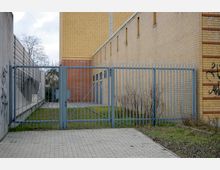 Gittertor, das einen kleinen gepflasterten Hof mit Rasen und Sträuchern abgrenzt, flankiert von Gebäudefassaden aus gelbem und rotem Backstein sowie einer Betonwand mit Graffiti. Im Hintergrund ist ein weiteres schmales Tor sichtbar, das zu einer weiteren Fläche führt.