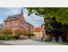 Innenhof mit Kopfsteinpflaster, umgeben von historischen Backsteingebäuden mit steilen Dächern und Spitzbogenfenstern. Im Vordergrund Bäume und Bänke, im rechten Bildbereich Glocken und eine Radfahrerin.