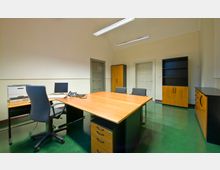 Büro mit glänzend grünem Boden, einem großen Holztisch, drei Stühlen, einem Schreibtischstuhl und Büromöbeln, darunter ein Drucker, ein Regal und Schränke. Im Hintergrund sind zwei graue Türen und Wandleuchten an der hohen, cremefarbenen Wand sichtbar.