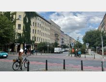 Straßenszene in einer Stadt mit mehrstöckigen Wohnhäusern im Altbaustil, teilweise mit Efeu bewachsen. Im Vordergrund eine Straße mit Fahrrad- und Fußgängerweg, mehreren Radfahrern, Autos und parkenden Fahrrädern; im Hintergrund Bäume und Straßencafés.