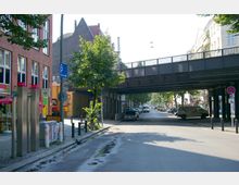 Straßenszene mit einer roten Backsteinfassade links und einer erhöhten schwarzen Eisenbahnbrücke, die die Straße überspannt. Im Vordergrund sind Gehwege mit Fahrradständern und geparkten Autos sowie ein Supermarkt sichtbar.