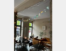 Gemütliches Café mit modernen schwarzen Tischen und Holzstühlen, beige Wänden und großen Fenstern, die viel Tageslicht hereinlassen. Dekorationen umfassen bunte Wimpelketten, Lichterketten, eine Zimmerpflanze und gerahmte Bilder an den Wänden.