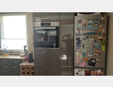 Moderne Küchenzeile mit Fenster über der Arbeitsplatte, Einbau-Backofen in einem Hochschrank und angrenzendem Edelstahlkühlschrank. Der Kühlschrank ist dicht mit Souvenir-Magneten und kleinen Anhängern beklebt; darunter stehen Arbeitsgeräte und ein kleines Weinregal.