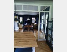 Wohnbereich mit offener Gestaltung, verbunden durch eine weiße Glastür. Im Vordergrund eine Holzarbeitsfläche, dahinter ein Essbereich mit Holztisch und Stühlen, sowie ein Wohnzimmer mit blauen Wänden, hellen Sesseln, einem Kamin und einem Fernseher. Der Raum hat einen Boden aus Fischgrätparkett und große Fenster mit Vorhängen.