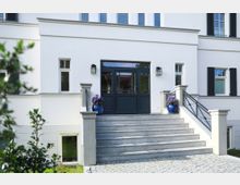 Weiße Hausfassade mit einer zentralen, modernen Eingangstür aus dunklem Holz und Glasfenstern. Eine breite Steintreppe mit Geländer führt zum Eingang, flankiert von blauen Blumentöpfen mit violetten Blumen.