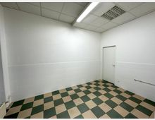 Leerer Raum mit weißen Wänden, teilweise gefliest, und grün-beigem Schachbrettmuster auf dem Boden. Eine weiße Tür befindet sich an der rechten Wand, die Decke hat Rasterpaneele und eine Leuchtstofflampe.