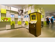 Ein modernes Museum mit einer Ausstellung über Videospielgeschichte. Im Vordergrund steht ein klassischer "Pong"-Spielautomat, während im Hintergrund Besucher interaktive Displays und Retro-Konsolen betrachten; die Wände sind mit farbigen Quadraten, Bildschirmen und Kopfhörern gestaltet.