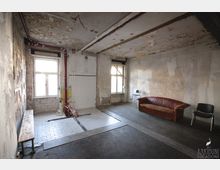 Ein roher, renovierungsbedürftiger Raum mit abblätternden Wänden und Sicht auf freigelegtes Mauerwerk. Der Raum enthält eine braune Ledercouch, zwei Stühle, eine Holzbänklein sowie gefliesten Bodenbereich mit sichtbaren Rohren und einem beschädigten Bereich.