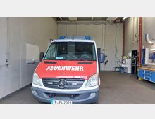 Ein Feuerwehrfahrzeug in einer Fahrzeughalle, umgeben von grauen Wänden, Werkzeugschränken und technischer Ausrüstung an den Seiten. Die Vorderseite des Fahrzeugs zeigt die Aufschrift „FEUERWEHR“ auf rotem Hintergrund.